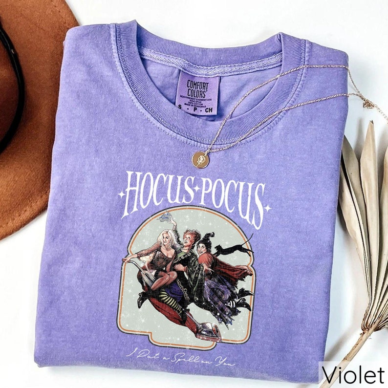 Puede incluir: Una camiseta violeta con las palabras "Hocus Pocus" en una fuente estilizada, con un gr&aacute;fico de tres brujas en una escoba. La frase "I Put a Spell on You" est&aacute; debajo del gr&aacute;fico. Un collar dorado con un colgante est&aacute; sobre la camiseta.