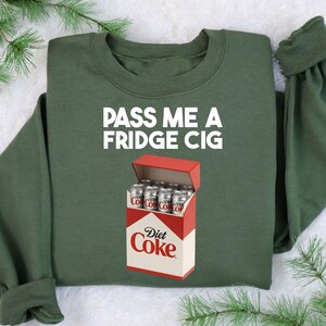 May include: An olive green sweatshirt with the text "PASS ME A FRIDGE CIG" in white. Below the text is a graphic of a Diet Coke box with several Diet Coke cans inside.