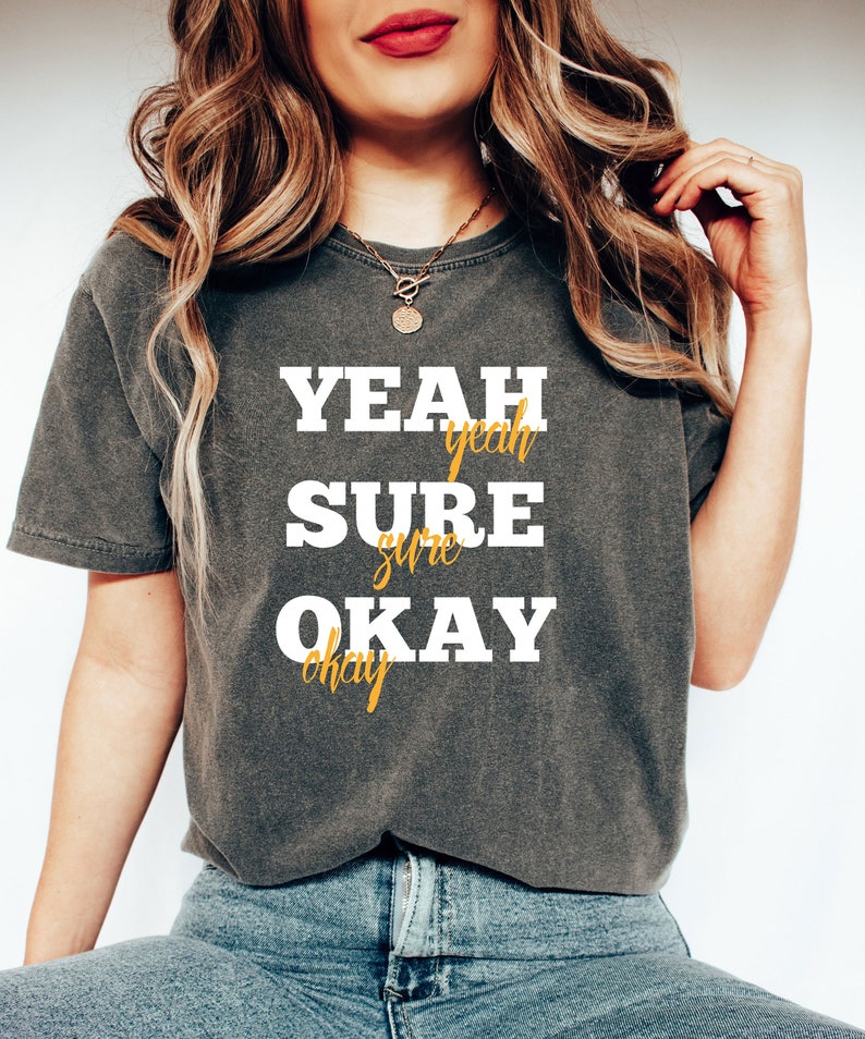 May include: A dark gray t-shirt with the text "YEAH SURE OKAY" in white letters. The words "yeah", "sure", and "okay" are written in a smaller font in yellow.