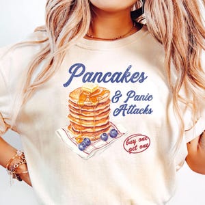 May include: A white t-shirt with a graphic of a stack of pancakes with syrup and butter, and the text "Pancakes & Panic Attacks" in blue. The text "buy one get one" is also included in red.