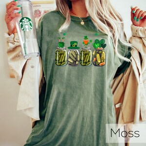 May include: A green t-shirt with a graphic of four pickle jars with leprechauns on top. The text "Moss" is visible in the bottom right corner of the image.