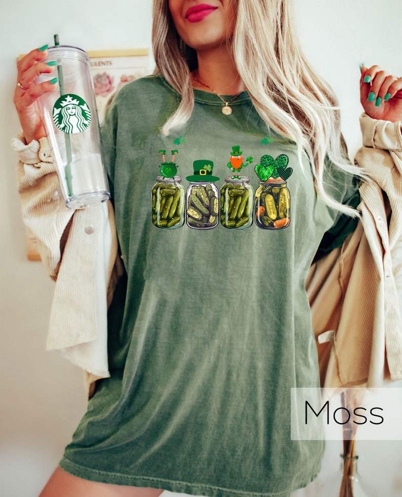 May include: A green t-shirt with a graphic of four pickle jars with leprechauns on top. The text "Moss" is visible in the bottom right corner of the image.