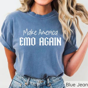 May include: A blue t-shirt with the white text "Make America EMO AGAIN." The shirt has short sleeves and a crew neck. The model is wearing blue jeans.