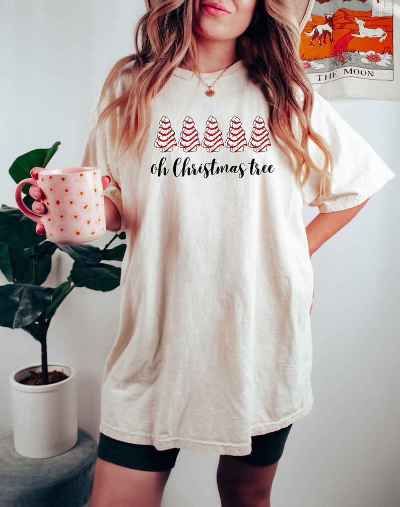 May include: Cream-colored t-shirt featuring a row of stylized red and white Christmas trees above the phrase "oh Christmas tree" in black script. The shirt is worn by a person holding a pink mug with white polka dots.