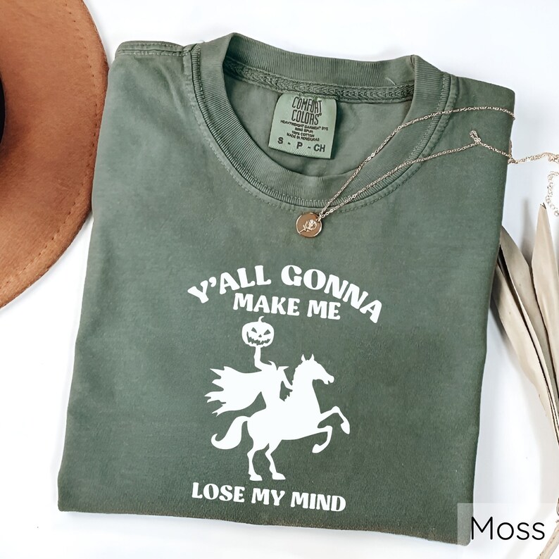 May include: A green t-shirt with a white graphic of a headless horseman riding a horse with a pumpkin for a head. The text on the shirt reads "Y'all gonna make me lose my mind". The shirt is labeled "Moss".