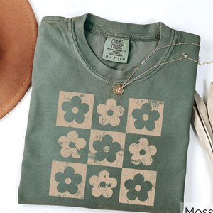 May include: A green t-shirt featuring a vintage-style design of nine brown flowers arranged in a grid pattern. The shirt has a tag that reads "Comfort Colors" and "S - P - Ch".