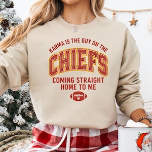 May include: A beige sweatshirt with the text "Karma is the Guy on the Chiefs Coming Straight Home to Me" in red and gold. A football graphic is below the text. The sweatshirt is part of a clothing collection.