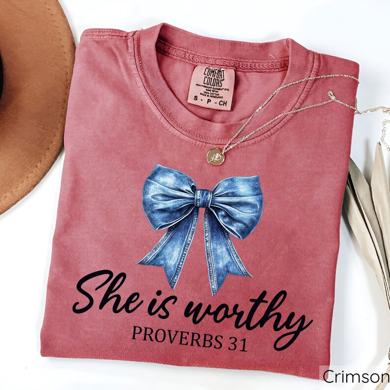 May include: A crimson red t-shirt with a blue denim bow and the text "She is worthy Proverbs 31" printed on the front.