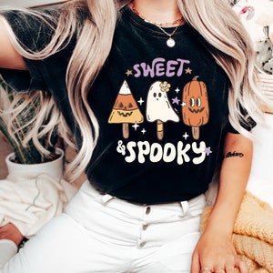 May include: Black t-shirt with a Halloween-themed graphic. The design features three popsicle-shaped characters: a candy corn, a ghost with a flower, and a pumpkin. The words "SWEET & SPOOKY" are written above and below the characters in a playful font.