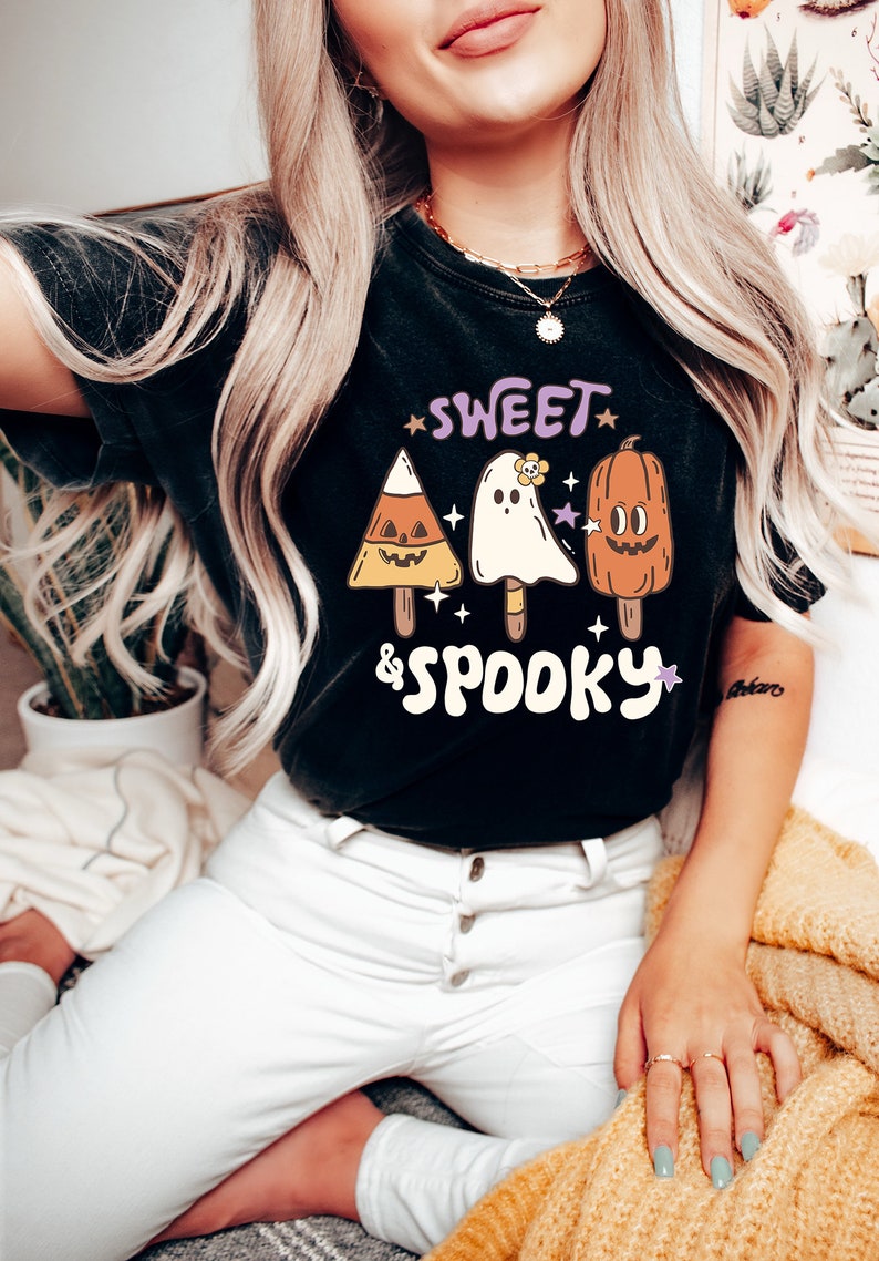 May include: Black t-shirt with a Halloween-themed graphic. The design features three popsicle-shaped characters: a candy corn, a ghost with a flower, and a pumpkin. The words "SWEET & SPOOKY" are written above and below the characters in a playful font.