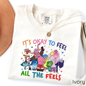 May include: A white t-shirt with a colorful graphic of characters from the movie Inside Out. The text "It's okay to feel all the feels" is printed on the shirt.