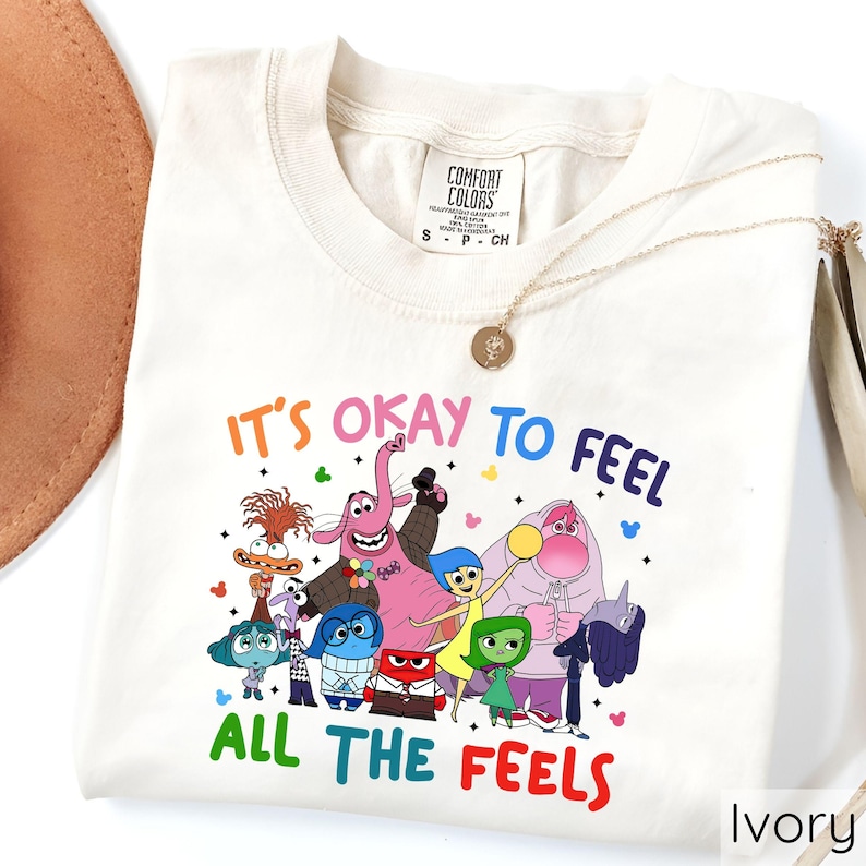 May include: A white t-shirt with a colorful graphic of characters from the movie Inside Out. The text "It's okay to feel all the feels" is printed on the shirt.
