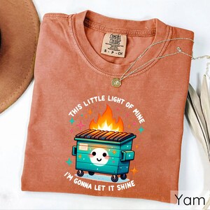 May include: A rust-colored t-shirt with a cartoon image of a green dumpster on fire. The text "This little light of mine" is above the dumpster and "I'm gonna let it shine" is below the dumpster.
