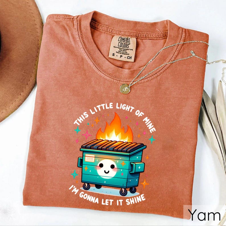 May include: A rust-colored t-shirt with a cartoon image of a green dumpster on fire. The text "This little light of mine" is above the dumpster and "I'm gonna let it shine" is below the dumpster.