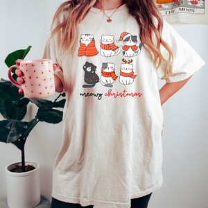 May include: An off-white t-shirt featuring a graphic of six cartoon cats wearing red and white winter accessories, including scarves and a Santa hat. The text below the cats reads meowy christmas in black and red cursive lettering.