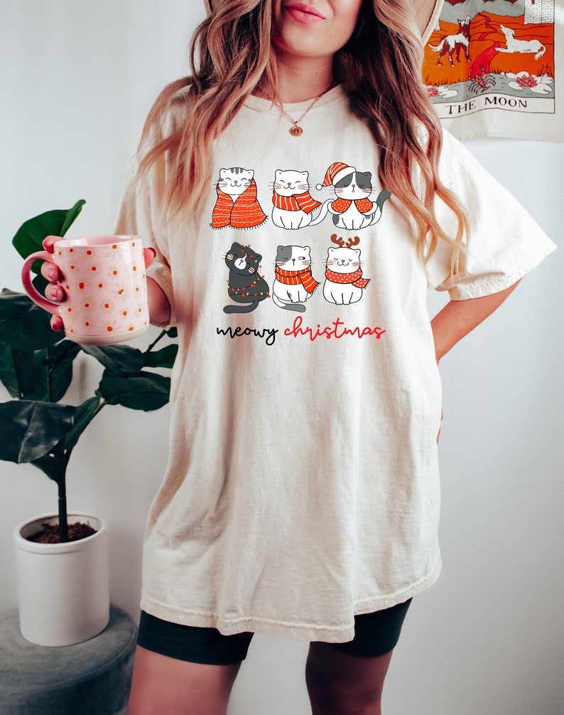 May include: An off-white t-shirt featuring a graphic of six cartoon cats wearing red and white winter accessories, including scarves and a Santa hat. The text below the cats reads meowy christmas in black and red cursive lettering.
