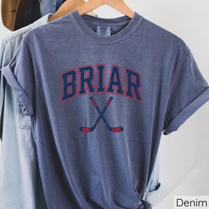 May include: A denim blue short-sleeve t-shirt featuring the word BRIAR in a dark blue arched font with a red outline. Below the text are two crossed hockey sticks in dark blue with red accents. The shirt has a crew neckline and a visible label.
