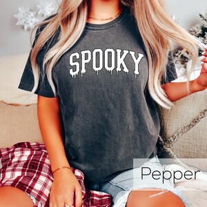 May include: Dark gray t-shirt with the word "SPOOKY" in white, dripping font. The shirt is a short-sleeved, crew neck style. The word "Pepper" is written in white in the bottom right corner of the image.