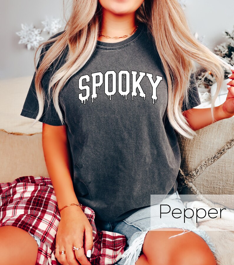 May include: Dark gray t-shirt with the word "SPOOKY" in white, dripping font. The shirt is a short-sleeved, crew neck style. The word "Pepper" is written in white in the bottom right corner of the image.