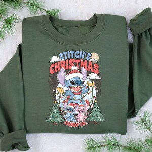 May include: Green sweatshirt featuring a graphic of Stitch in a Santa hat holding Christmas lights. The text "Stitch's Christmas Candy Co." is displayed above and below the character. Two smaller Stitch characters are also wearing Santa hats.
