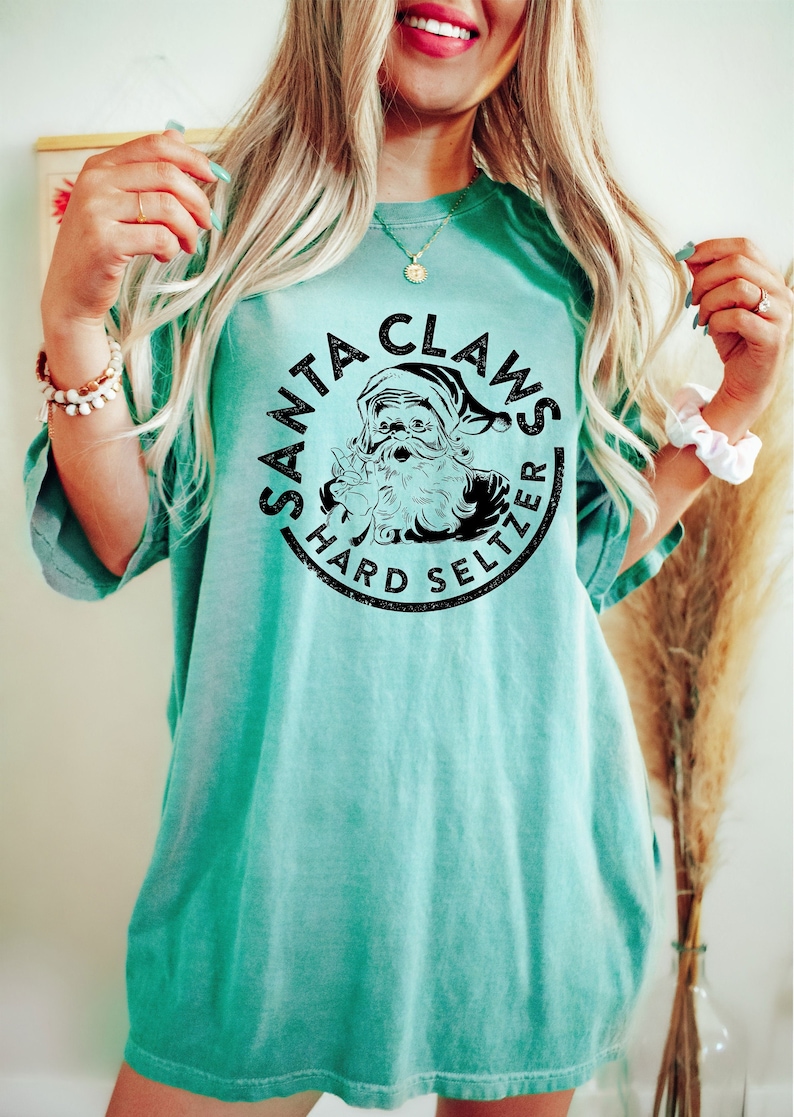 May include: A turquoise t-shirt with a black graphic that reads "SANTA CLAWS HARD SELTZER" in a circular design. The graphic features a Santa Claus illustration. The shirt has short sleeves and a relaxed fit.