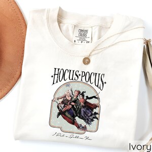 Puede incluir: Camiseta de color marfil con la frase "Hocus Pocus" y un gr&aacute;fico de tres brujas volando en una escoba. La camiseta tambi&eacute;n tiene el texto "I Put a Spell on You". Un collar dorado con un colgante est&aacute; sobre la camiseta.