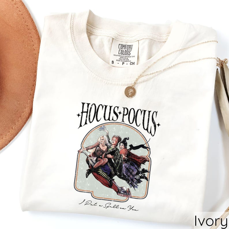 Puede incluir: Camiseta de color marfil con la frase "Hocus Pocus" y un gr&aacute;fico de tres brujas volando en una escoba. La camiseta tambi&eacute;n tiene el texto "I Put a Spell on You". Un collar dorado con un colgante est&aacute; sobre la camiseta.