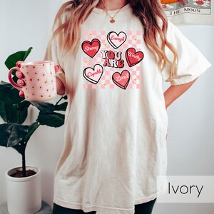 Puede incluir: Una mujer lleva una camiseta beige claro con un fondo a cuadros rosa y blanco y corazones rosas con afirmaciones positivas. El texto en los corazones dice "You Are", "Strong", "Capable", "Worthy", "Loved", y "Enough". La camiseta est&aacute; etiquetada como "Ivory".