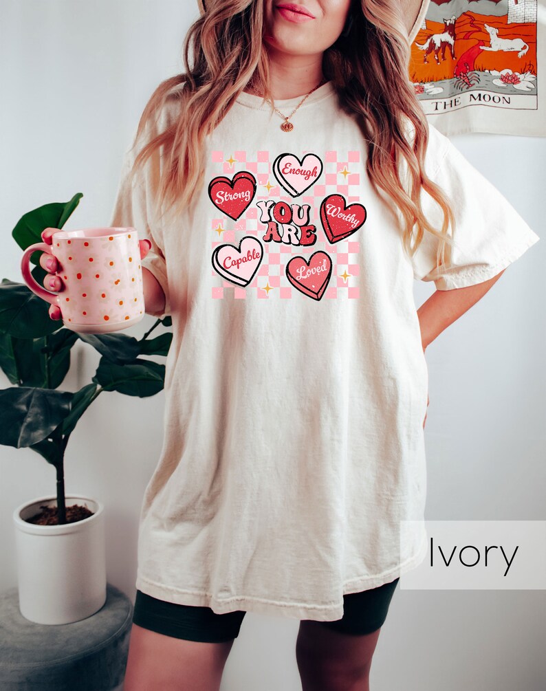 Puede incluir: Una mujer lleva una camiseta beige claro con un fondo a cuadros rosa y blanco y corazones rosas con afirmaciones positivas. El texto en los corazones dice "You Are", "Strong", "Capable", "Worthy", "Loved", y "Enough". La camiseta est&aacute; etiquetada como "Ivory".