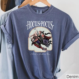 Puede incluir: Camiseta azul denim con la frase "Hocus Pocus" sobre una ilustraci&oacute;n de tres brujas en una escoba. La frase "I Put a Spell on You" est&aacute; impresa debajo. La camiseta est&aacute; colgada en una percha de madera.
