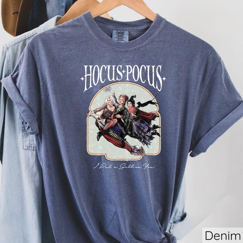 Puede incluir: Camiseta azul denim con la frase "Hocus Pocus" sobre una ilustraci&oacute;n de tres brujas en una escoba. La frase "I Put a Spell on You" est&aacute; impresa debajo. La camiseta est&aacute; colgada en una percha de madera.