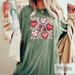 Puede incluir: Camiseta verde con fondo a cuadros rosa y blanco. El fondo tiene cinco corazones con las palabras "You Are" en el centro y "Strong", "Enough", "Worthy", "Capable" y "Loved" en los corazones. El texto "Moss" est&aacute; en la parte inferior de la imagen.