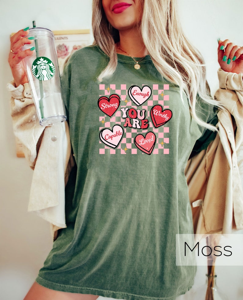 Puede incluir: Camiseta verde con fondo a cuadros rosa y blanco. El fondo tiene cinco corazones con las palabras "You Are" en el centro y "Strong", "Enough", "Worthy", "Capable" y "Loved" en los corazones. El texto "Moss" est&aacute; en la parte inferior de la imagen.
