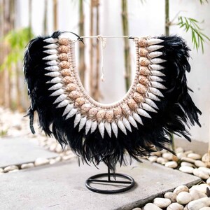 Peut inclure: Collier artisanal composé de plumes noires, de coquillages blancs et d'éléments beiges. Le collier est présenté sur un support en métal noir, avec des tiges de bambou et des galets blancs en arrière-plan.