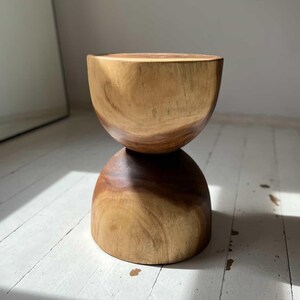 Op de afbeelding: Een houten kruk met een unieke zandlopervorm. De kruk is gemaakt van twee gestapelde, afgeronde stukken hout, waardoor een glad, organisch ontwerp ontstaat.