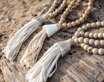 May include: Three wooden bead necklaces with white tassel ends. The beads are natural wood and the tassels are made of cotton. The necklaces are displayed on a piece of driftwood.
