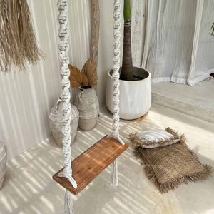 Op de afbeelding: Een witte macramé schommel met een houten zitting hangt aan een houten pergola. De schommel bevindt zich in een kamer met witte muren en een plant in een pot.