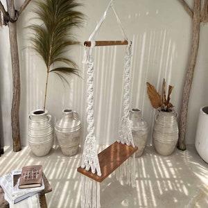 Könnte beinhalten: Weiße Makrame-Schaukel mit einem Holz-Sitz, die an einer Holzpergola hängt. Die Schaukel befindet sich in einem weißen Raum mit Topfpflanzen und einem Buch auf einem Tisch.