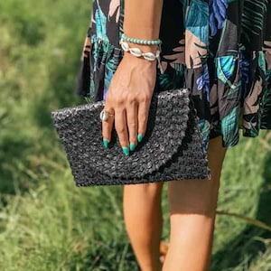 Może przedstawiać: Kobieta w czarnej i zielonej sukience z tropikalnym nadrukiem, z czarną plecioną kopertówką i bransoletką z muszli.