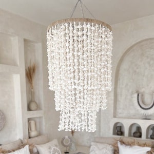 Op de afbeelding: Een witte, met schelpen versierde kroonluchter hangt boven een witte hoekbank met witte en beige kussens. Een houten salontafel met twee houten kaarsenstandaards staat voor de bank. Een houten kruk staat op de voorgrond.