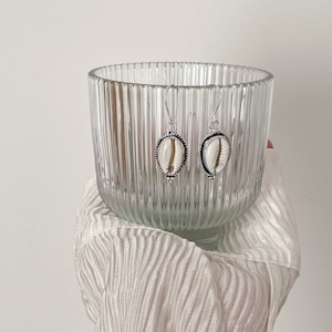 May include: A pair of silver earrings with white cowrie shell accents. The earrings are hanging from a glass bowl.