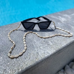 May include: Black sunglasses with a seashell chain. The sunglasses are cat-eye shaped. The chain is made of small, cream-colored seashells. The background shows a blue swimming pool.