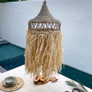 May include: A woven pendant light with a fringe made of natural fibers. The light is hanging over a table set with plates, bowls, and candles.