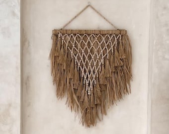 Puede incluir: Un tapiz de macramé triangular hecho de cuerda color canela con cuentas blancas dispuestas en patrón de diamante, con largos flecos en la parte inferior. Al lado hay una decoración de fibras marrones en un jarrón de cerámica blanco.
