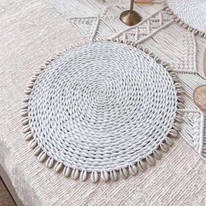 May include: White woven placemats with a beaded edge, set on a white tablecloth with a macrame runner. There are candlesticks with white candles and glasses on the table.
