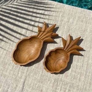 May include: Two wooden pineapple-shaped bowls, one larger than the other, are displayed on a light beige textured surface. The bowls are carved from light brown wood and have a natural, rustic finish.