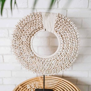 Puede incluir: Un collar de conchas blancas sobre un soporte negro. El collar está hecho de pequeñas conchas blancas que están dispuestas en un patrón circular. El soporte está hecho de metal y tiene una base cuadrada.