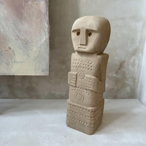 Peut inclure: Statue en bois sculpté représentant une figure avec une tête arrondie et un corps rectangulaire. La statue marron clair présente des sculptures détaillées sur la poitrine et le bas du corps. Objet décoratif fait à la main.