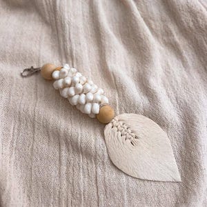 Peut inclure: Un porte-clés fait à la main avec un groupe de petites perles blanches, des perles en bois et un pendentif en forme de feuille, texturé, blanc cassé. Le porte-clés a un fermoir argenté pour une fixation facile. Le fond est un tissu beige clair.
