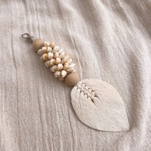 以下が含まれることがあります： 小さなクリーム色の貝殻と木製のビーズのクラスターが特徴の手作りのキーホルダー。 キーホルダーは、オフホワイトのテクスチャード加工された素材で作られた葉の形をしたペンダントと、シルバー色の留め金が付いています。